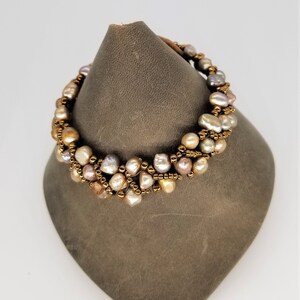 May include: A multi-strand bracelet featuring irregularly shaped pearls in shades of white, peach, and gray. The pearls are strung together with gold-colored beads and a brown leather cord.