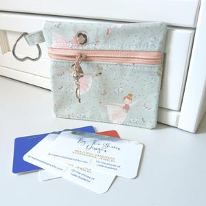 May include: A small, square coin purse with a light blue background and a pink zipper. The purse features a pattern of ballerinas and flowers. A silver key ring and a ballerina charm hang from the zipper. Business cards are in front of the purse.
