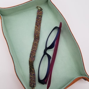 Puede incluir: Una bandeja de cuero verde claro con un brazalete de cuentas y un par de gafas en su interior.