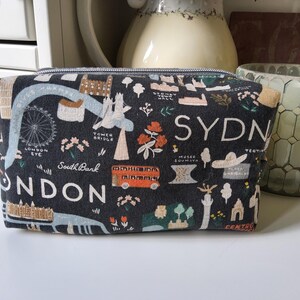 Puede incluir: Un estuche rectangular de tela con fondo gris oscuro y un estampado colorido de mapas de Londres y Sídney. El diseño incluye ilustraciones del London Eye, el Tower Bridge, un autobús rojo de dos pisos y etiquetas de texto como London, Sydney y South Bank.