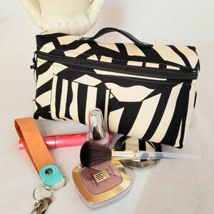 May include: Black and white striped zippered makeup bag with a black handle. The bag is open and there are makeup brushes, a compact, and nail polish inside.