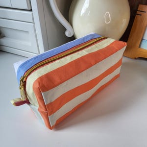 Puede incluir: Un estuche rectangular de tela con rayas horizontales naranjas y crema en el frente y los lados. El panel superior es azul claro y el cierre consiste en una cremallera de metal dorado. Una pequeña lengüeta de tela está unida al lado de la cremallera.