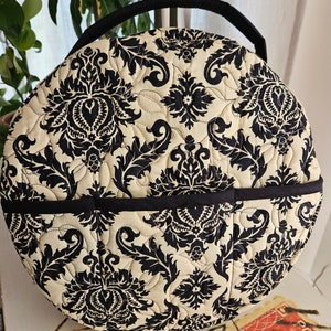 May include: A round, quilted storage bag with a black handle and black trim. The bag is covered in a cream and black damask pattern.