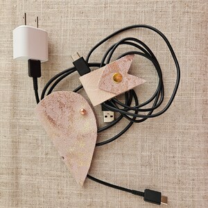 Puede incluir: Dos organizadores de cables de cuero en rosa y beige con detalles dorados. Los organizadores están envueltos alrededor de cables negros y un cargador de teléfono blanco.