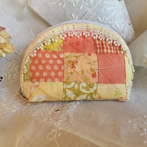 May include: A patchwork coin purse with a half-moon shape. The purse features a variety of fabrics in shades of yellow, peach, and green, with floral and geometric patterns. A white lace trim accents the zipper closure.