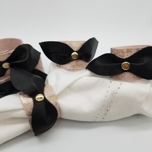 May include: Three pink and gold leather napkin rings with black leather bows and gold accents. The rings are tied around a white napkin.