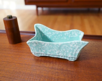 May include: A light blue ceramic planter with a white floral pattern. The planter has a unique, wavy shape and is sitting on a wooden surface.