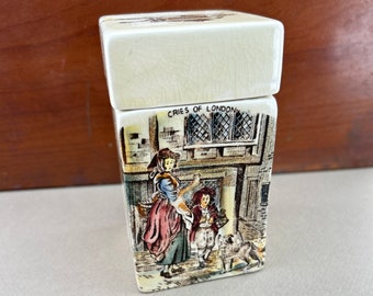May include: Cream-colored ceramic box with a lid, featuring a detailed illustration of a street scene with figures and the text "CRIES OF LONDON." The box has a rectangular shape and a slightly cracked glaze.