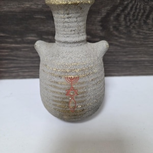 Puede incluir: Un pequeño jarrón gris texturizado con un borde dorado y rayas horizontales doradas. El jarrón tiene dos asas pequeñas y un diseño rojo de la Estrella de David y la menorá. El jarrón es probablemente de cerámica o alfarería.