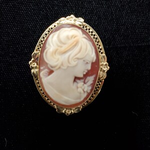 May include: A gold-toned cameo brooch featuring a carved portrait of a woman with a floral design. The cameo is set in an oval frame with a detailed design.