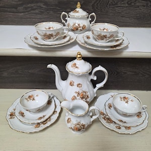 Puede incluir: Un juego de té vintage de porcelana blanca con un estampado floral de rosas en tonos marrón y dorado. El juego incluye una tetera, una lechera, una azucarera, tazas y platillos. Los bordes están adornados con oro.