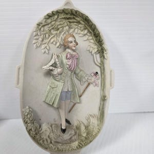 May include: Oval-shaped ceramic wall plaque featuring a relief of a man in 18th-century attire. The figure is in a light green coat, pink bow, and blue shorts, holding a flower and a walking stick. The background includes a tree and foliage.