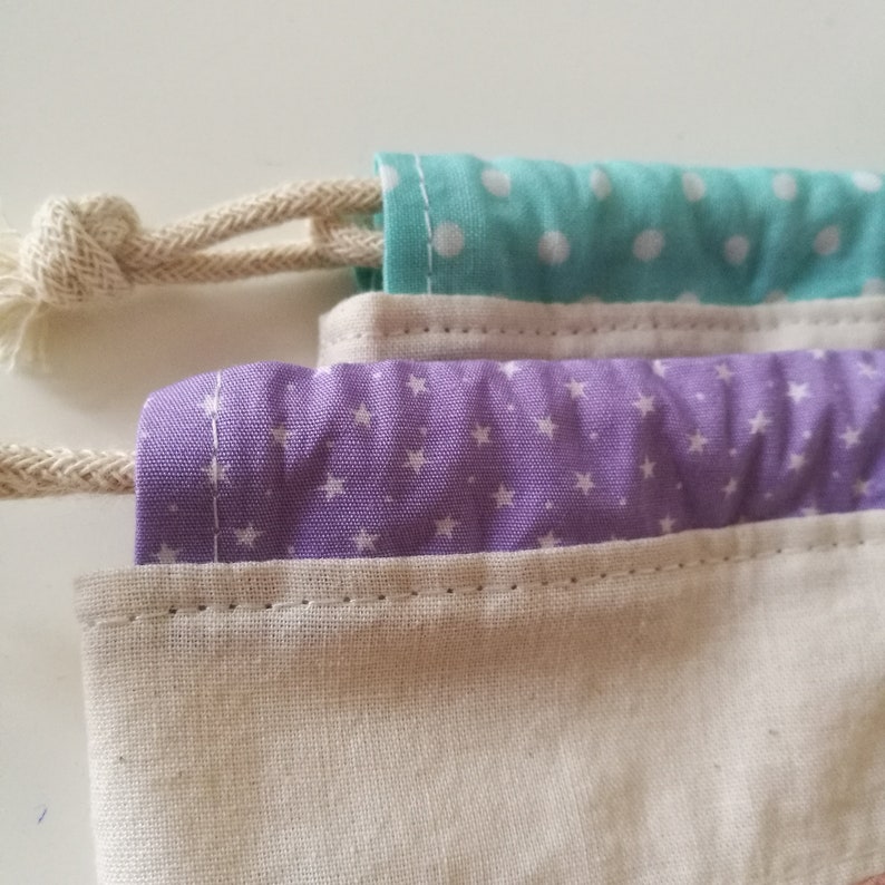 May include: A drawstring bag made of natural fabric with three layers. The top layer is a light blue fabric with white polka dots. The middle layer is a purple fabric with white stars. The bottom layer is a natural fabric.