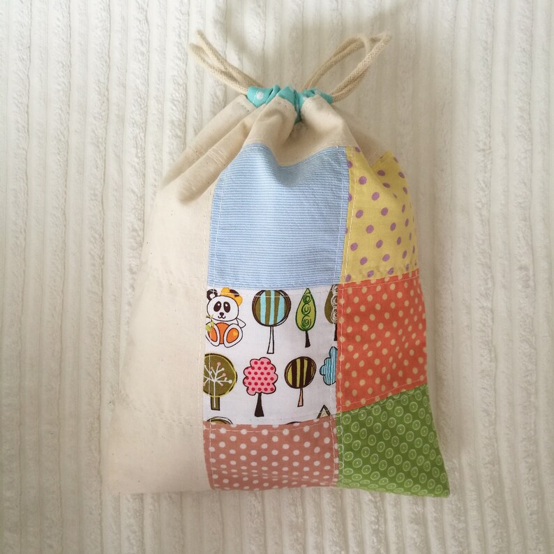 May include: A white canvas drawstring bag with a patchwork design. The patches are made of different fabrics with various patterns, including polka dots, stripes, and a panda bear design.