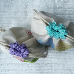 May include: Two drawstring bags made of natural fabric with a light blue and yellow patterned design. The bags have a purple and a turquoise ribbon tied around the top.
