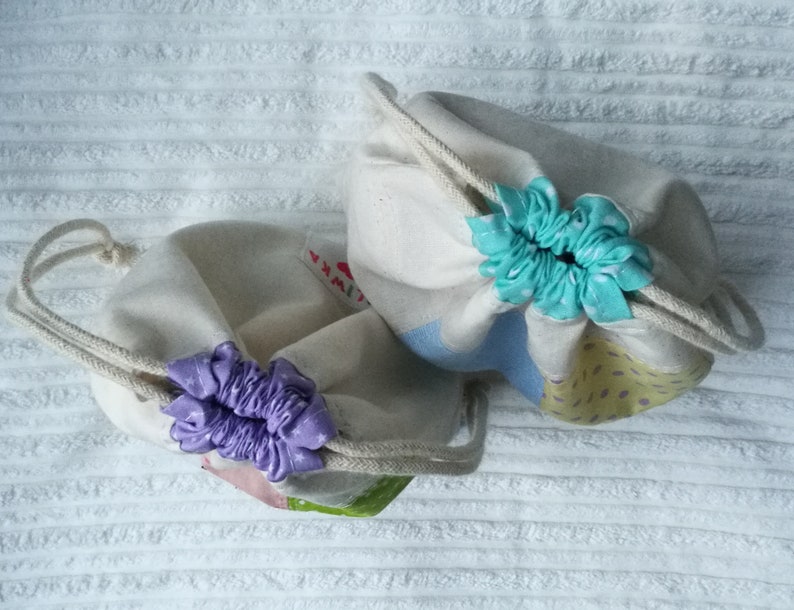 May include: Two drawstring bags made of natural fabric with a light blue and yellow patterned design. The bags have a purple and a turquoise ribbon tied around the top.