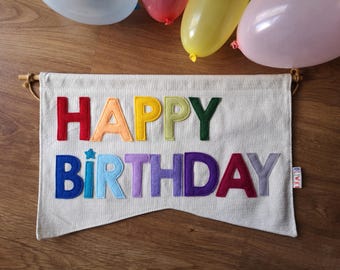 Może przedstawiać: Biały baner z napisem "Happy Birthday" w kolorach tęczy z filcu. Baner jest zawieszony na drewnianym drążku.