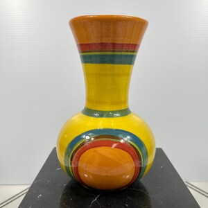May include: A colorful ceramic vase with a bulbous base and a flared top. The vase features a vibrant design with bands of orange, red, green, and yellow. The base has concentric circles of orange, red, and blue.