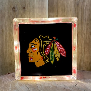 May include: A black light-up glass block with a white outline and a red, yellow, green, and black Chicago Blackhawks logo. The logo features a Native American profile with a headdress.