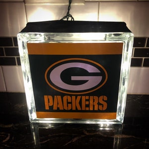 May include: A glass block with a black background and an orange and white Green Bay Packers logo. The block is lit from within with white LED lights.