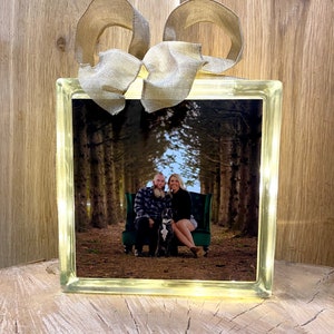 May include: A glass block with a photo of a couple and their dog sitting on a green velvet chair in a forest. The block is lit from behind and has a brown burlap ribbon tied around it.