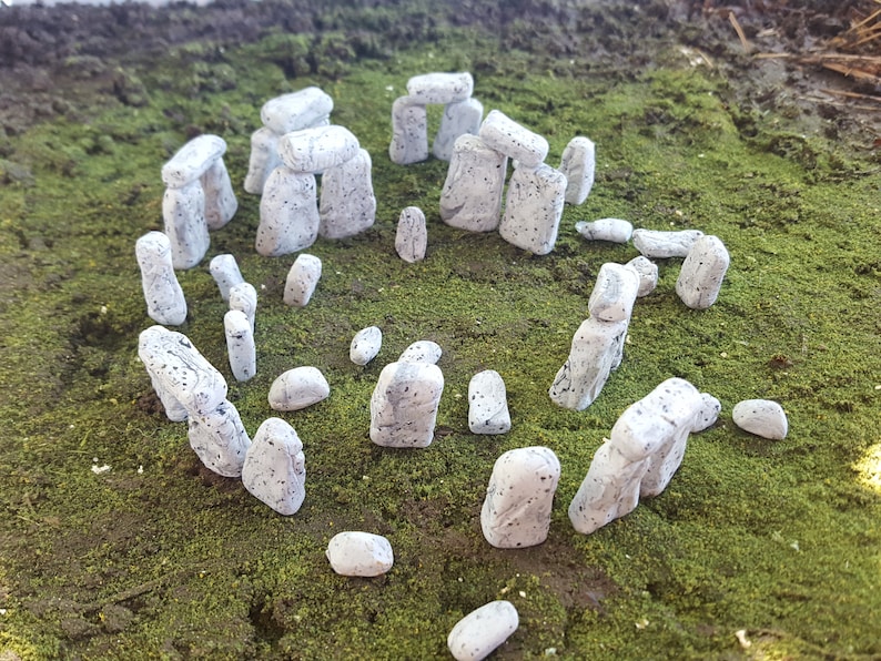 May include: A miniature model of Stonehenge made of white painted stones with black speckles. The stones are arranged in a circle on a bed of green moss.