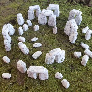 May include: A miniature model of Stonehenge, a prehistoric monument in England, made of white painted stones. The model is made up of several arches and individual stones, all arranged in a circle on a green moss surface.