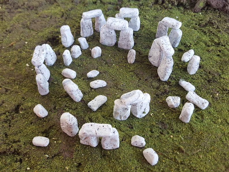 May include: A miniature model of Stonehenge, a prehistoric monument in England, made of white painted stones. The model is made up of several arches and individual stones, all arranged in a circle on a green moss surface.