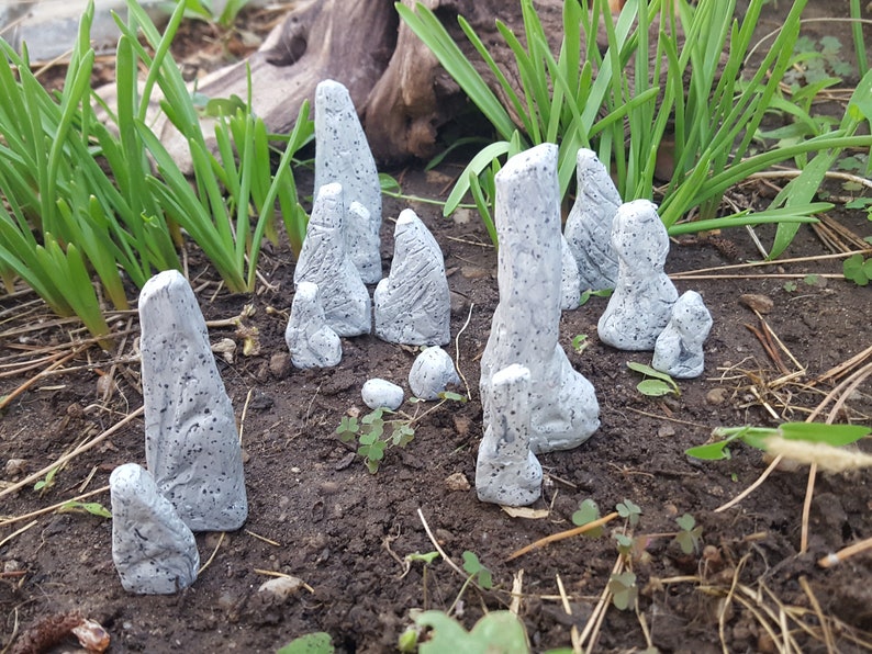 Może przedstawiać: Kolekcja małych, szarych, teksturowanych figurek przypominających kamienie, ułożonych na brązowej ziemi. Figurki mają kształt wysokich, cienkich filar&oacute;w i są rozrzucone po całym obrazie.