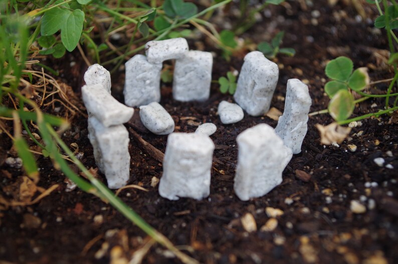 May include: A miniature Stonehenge replica made of white painted foam, arranged in a circle on a bed of brown soil. The miniature Stonehenge is surrounded by green plants.