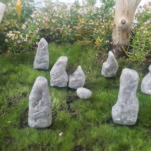 Może przedstawiać: Zestaw ośmiu miniaturowych figurek z szarego kamienia ułożonych na zielonym mchu. Figurki mają r&oacute;żne kształty i rozmiary, przypominając małe stojące kamienie.