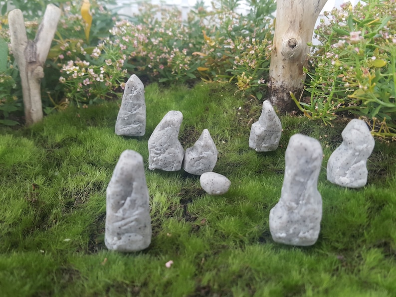 Może przedstawiać: Zestaw ośmiu miniaturowych figurek z szarego kamienia ułożonych na zielonym mchu. Figurki mają r&oacute;żne kształty i rozmiary, przypominając małe stojące kamienie.