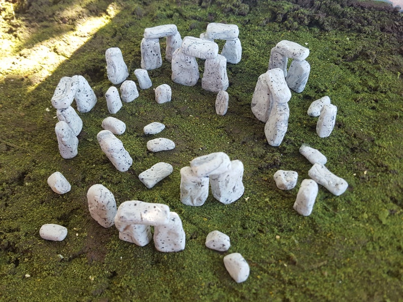 May include: A miniature model of Stonehenge made of white stone with a speckled texture. The model is set on a bed of green moss.