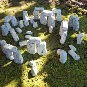 May include: A miniature Stonehenge model made of grey stones arranged in a circle on a green mossy surface.