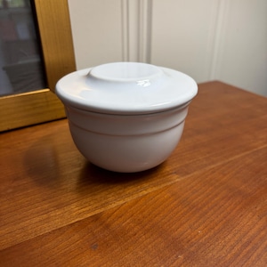 May include: A white ceramic bowl with a matching lid. The bowl features a smooth, glossy finish and a rounded base, while the lid has a slightly raised circular center. The design is simple and functional, with a clean, uniform white color throughout.