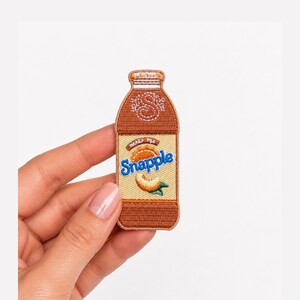 May include: An embroidered patch shaped like a Snapple bottle with a brown cap and body, featuring a tan label with the blue text Snapple, a small orange peach graphic, and a white letter S stitched on the upper portion of the bottle design.