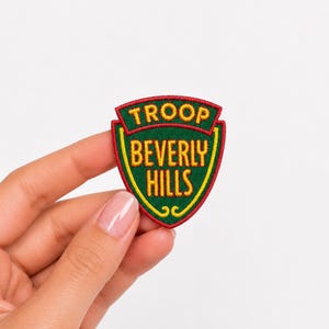 May include: A shield-shaped embroidered patch featuring a dark green background with a red border and yellow trim. The text TROOP BEVERLY HILLS is stitched in yellow thread across the center of the patch in a bold, sans-serif font style.