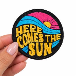 May include: A circular embroidered patch with a black border featuring the yellow text "HERE COMES THE SUN" layered over a design of blue waves and a pink sky with a yellow sun rising in the upper right corner of the circular fabric patch.