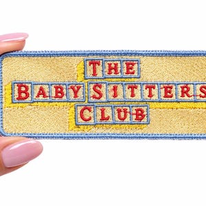 May include: A rectangular embroidered patch with a light yellow background and a blue border. The text THE BABY-SITTERS CLUB is stitched in red, with each letter enclosed in individual blue-outlined yellow squares arranged in three rows.