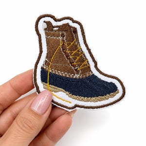 May include: An embroidered patch shaped like a duck boot with a brown upper, a dark navy blue rubberized lower section, and yellow laces. The patch features a thick white border outline and detailed stitching throughout the design.