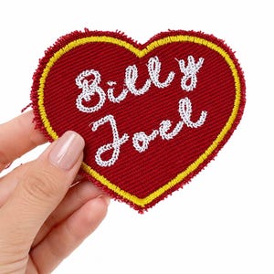 May include: A heart-shaped embroidered patch featuring a deep red fabric base with a bright yellow border. The text "Billy Joel" is stitched in white cursive thread across the center of the heart, which has a textured, fabric-like appearance.