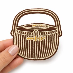 May include: An embroidered patch shaped like a round basket with a handle. The design features dark brown and cream vertical stripes, a solid dark brown lid, and a small gold-colored latch detail centered on the front of the basket body.