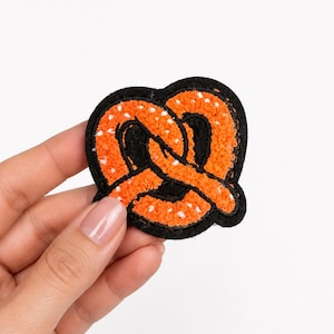 May include: A pretzel-shaped patch featuring a textured orange surface with small white dots, outlined by a thick black border. The design is crafted with a fuzzy, raised material that mimics the appearance of a baked dough snack.