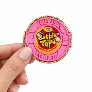 May include: A circular embroidered patch featuring a pink and yellow design with the text Bubble Tape and Awesome Original. The patch has a gold glitter border and depicts a stylized graphic of bubble gum in a round container with a pink lid.