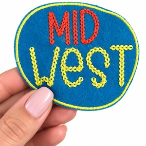 May include: A circular blue felt patch featuring the word MID embroidered in red thread and the word WEST embroidered in yellow thread. The patch has a yellow embroidered border around the entire outer edge of the circular blue felt shape.