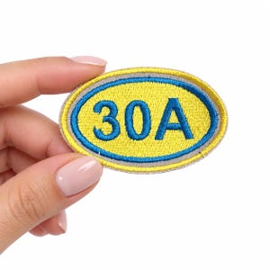 May include: An oval-shaped embroidered patch featuring a bright yellow center with the blue text 30A. The patch is bordered by a thin blue ring and an outer light gray edge, creating a distinct layered design suitable for attachment to fabric items.