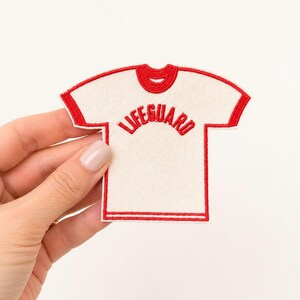 May include: An embroidered patch shaped like a t-shirt with a cream-colored base and bright red trim along the neckline, sleeves, and hem. The word LIFEGUARD is stitched in red capital letters across the chest in a curved, arched orientation.