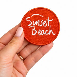 May include: A circular embroidered patch with a vibrant orange background and a stitched border. The text Sunset Beach is embroidered in white cursive lettering across the center of the patch, featuring a small curved line above the word Sunset.