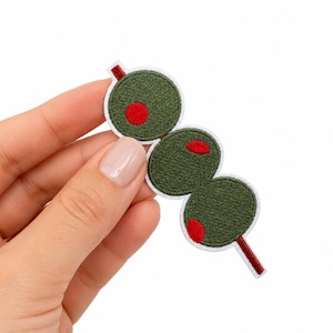 May include: An embroidered patch featuring three round, olive-green circles arranged in a vertical line, each with a small, bright red circular center, all pierced by a thin, dark red line representing a cocktail pick with a white border.