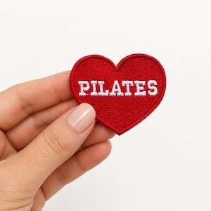May include: A heart-shaped embroidered patch in a vibrant red color featuring the word PILATES stitched in white capital letters across the center. The patch has a finished border and a textured fabric surface, designed for application to textiles.